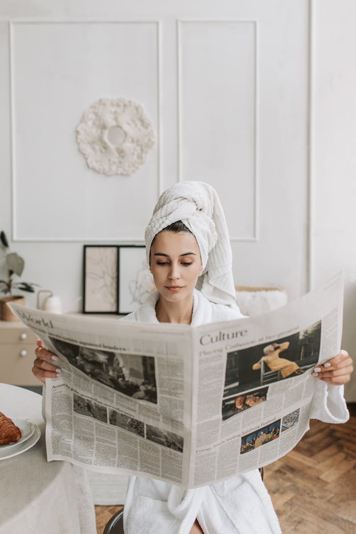 Woman in a bathrobe with head towel reading a newspaper, enjoying a quiet morning indoors.